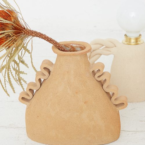 A hand-made ceramic vase with a ruffled design holding dried plants, accompanied by a Clandestine Amphora Soleil vase in Raw Sunny Brown Clay with a bulbous body in the background.